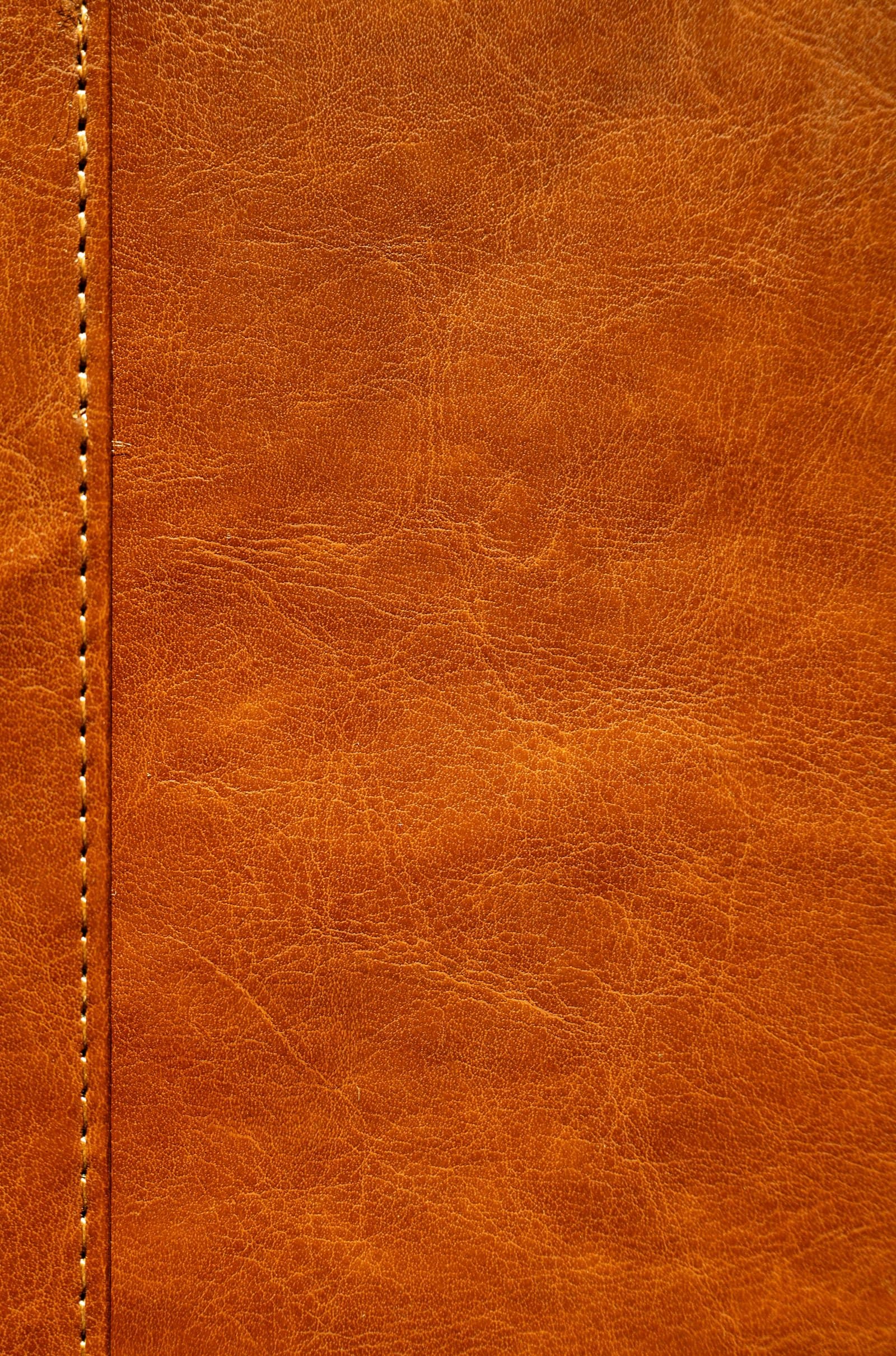 Smooth tan full-grain leather beside a hand-stitched seam, natural growth lines and open pores visible under a low sideways light