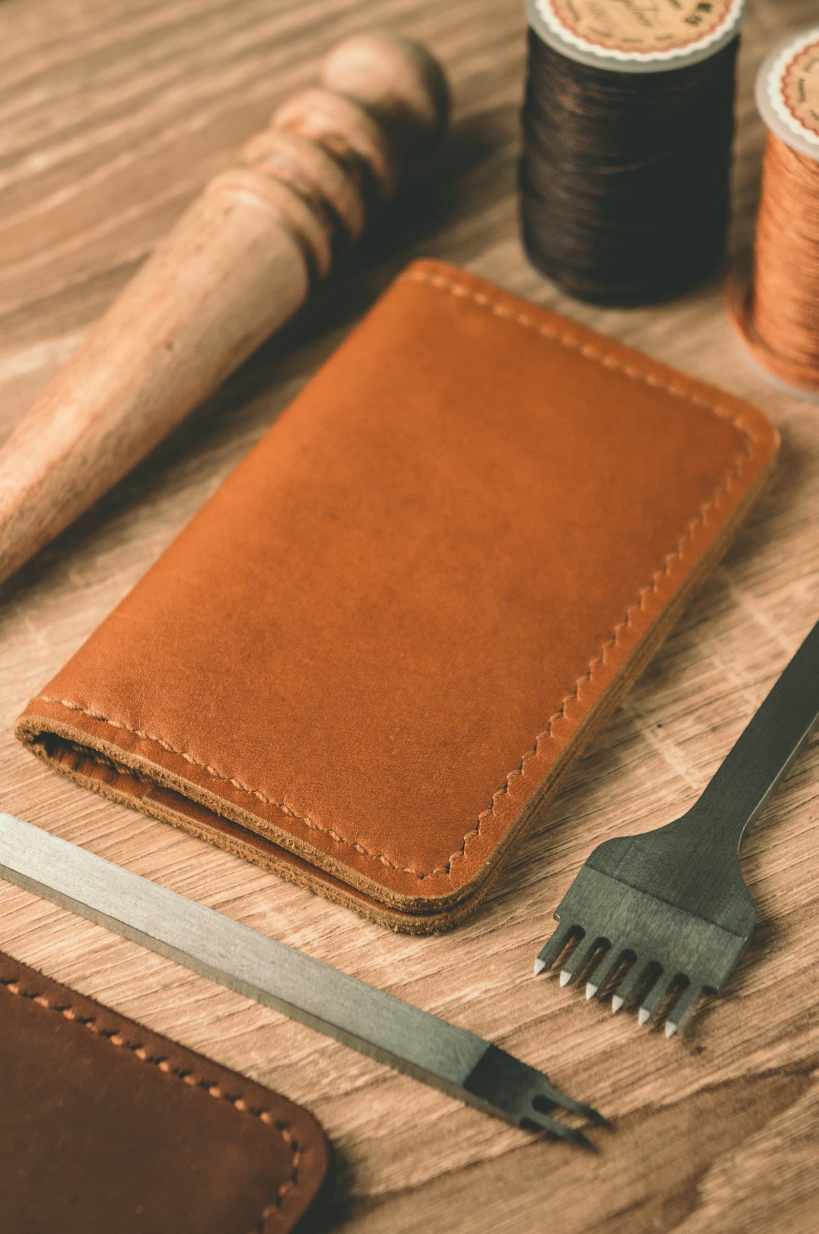 A natural undyed leather cardholder on a leatherworker's bench beside waxed thread and a stitching iron