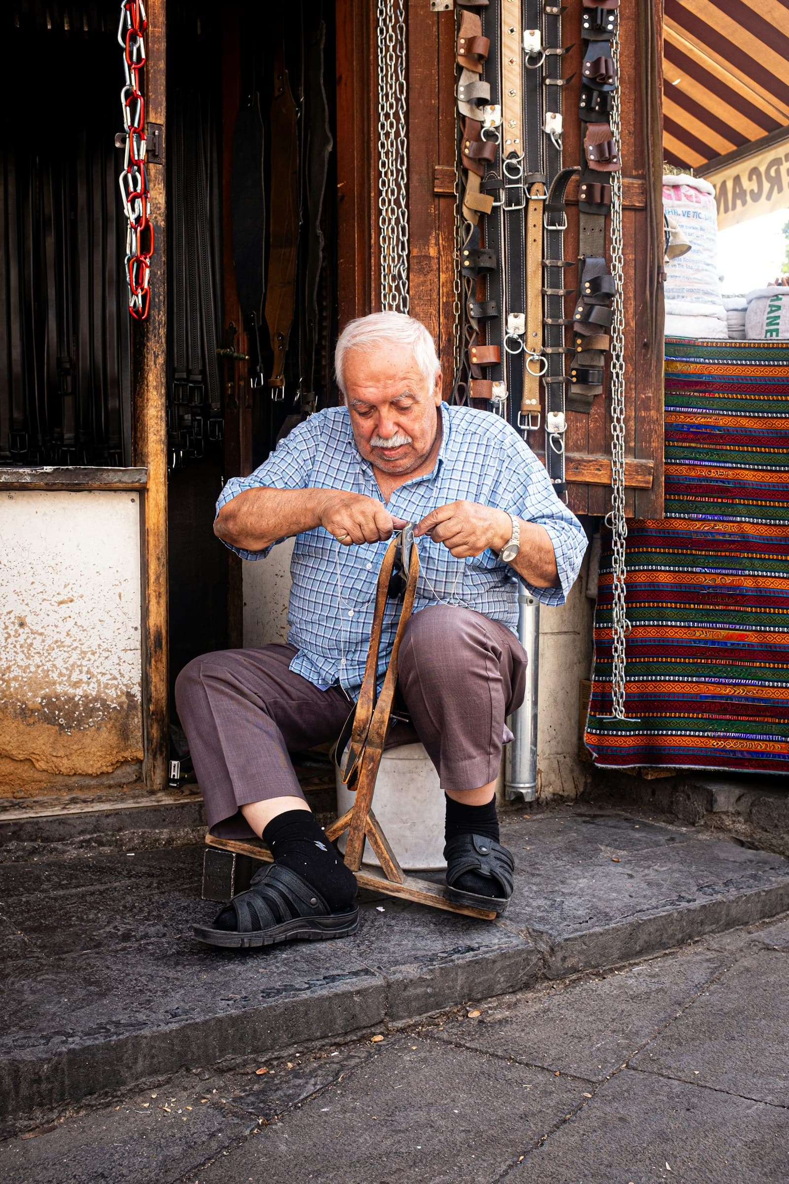 A leatherworker drawing a strap taut across his knee in the doorway of his workshop